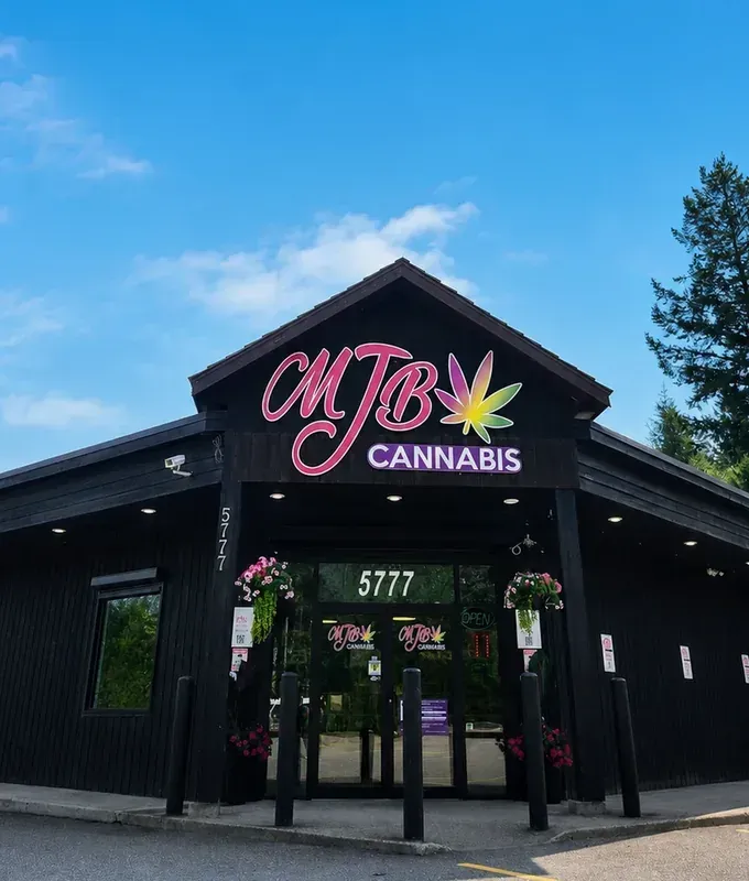 The MJB Cannabis storefront at 5777 Albert Place: a black wood building with the pink MJB Cannabis sign, hanging flower baskets and tall evergreens under a blue sky.