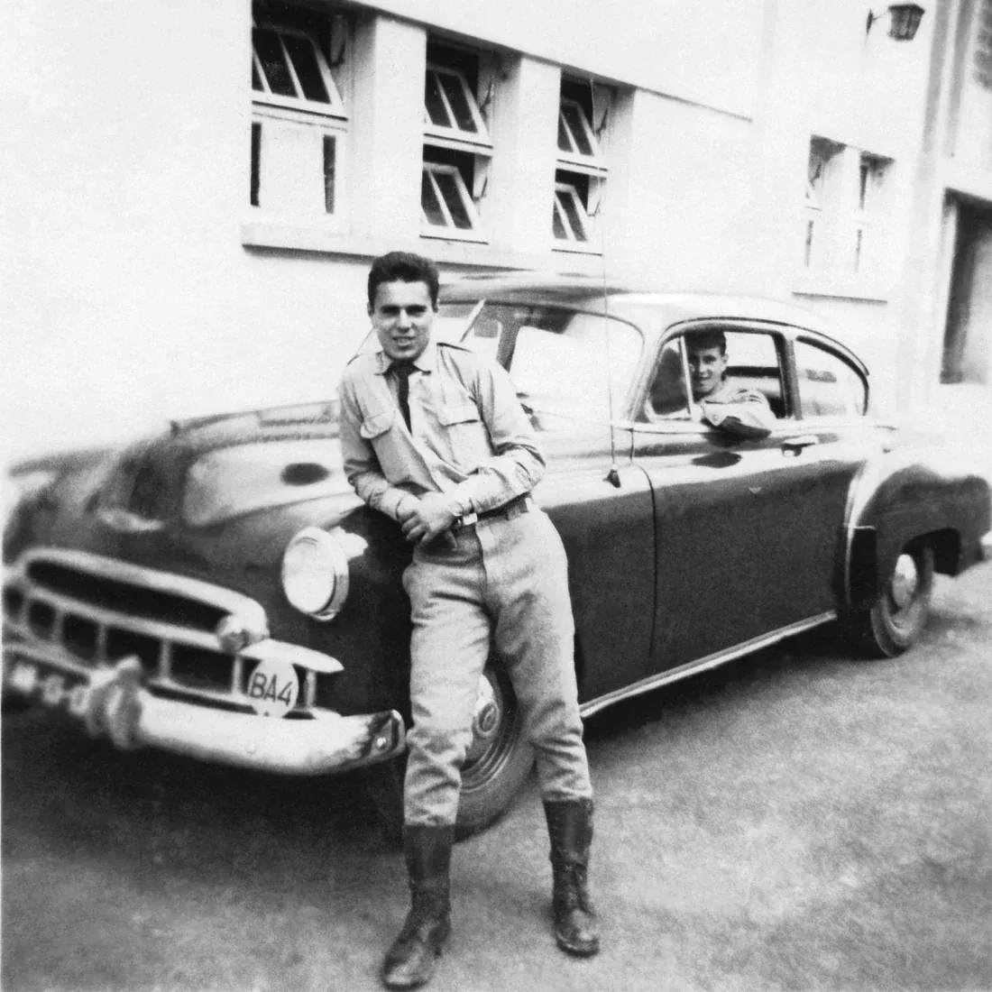 Black-and-white photo of a young man in a pressed shirt and tall boots, smiling as he leans on a 1950s car, while a friend grins from the driver’s window.