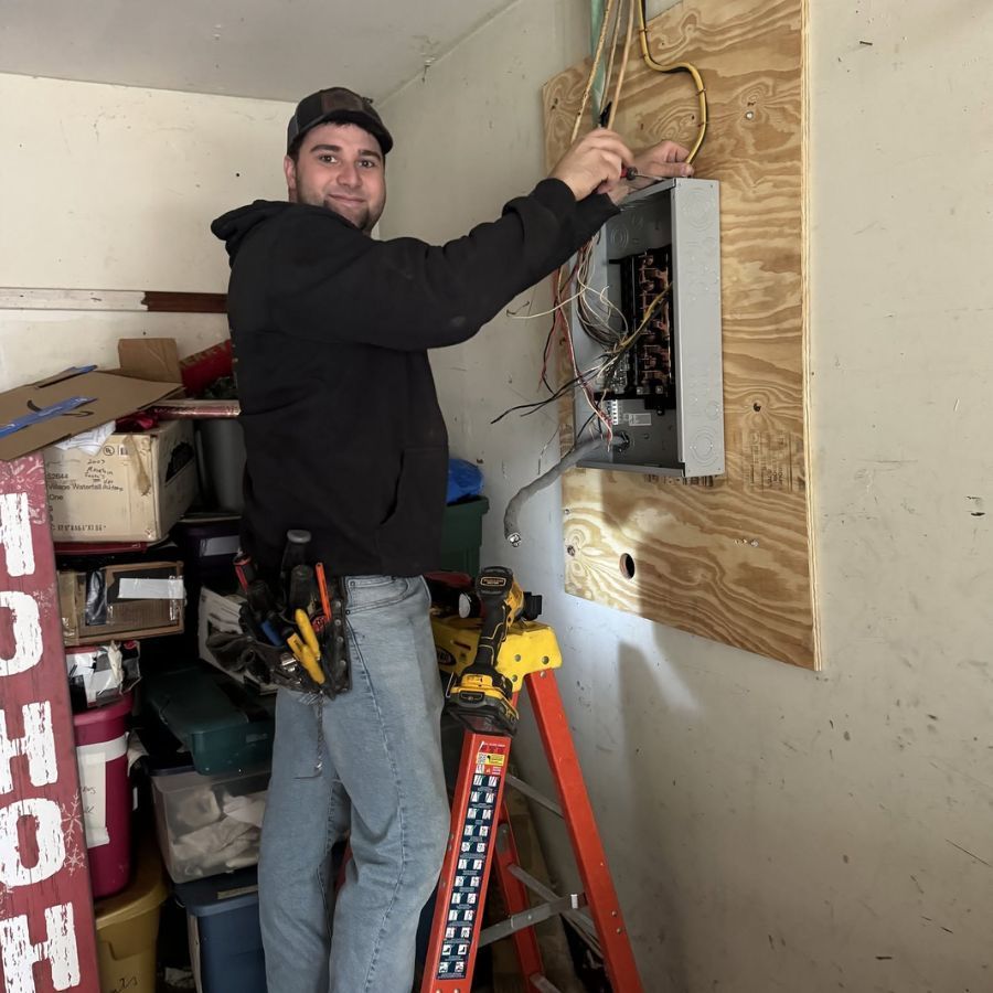 Technician wiring an electrical panel mounted on plywood in a garage-like room, using a ladder.