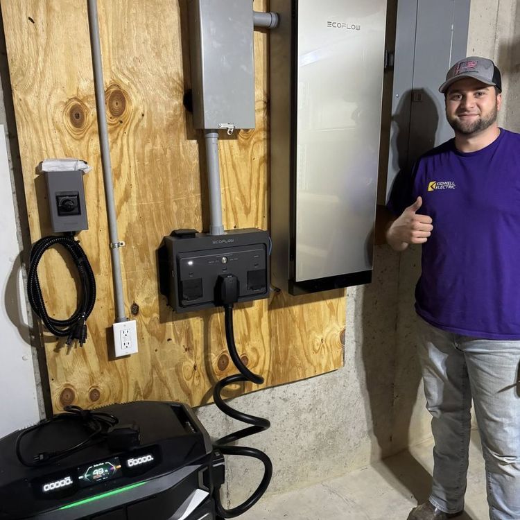 Technician beside wall-mounted electrical panel and outlet, giving a thumbs-up in a workshop.