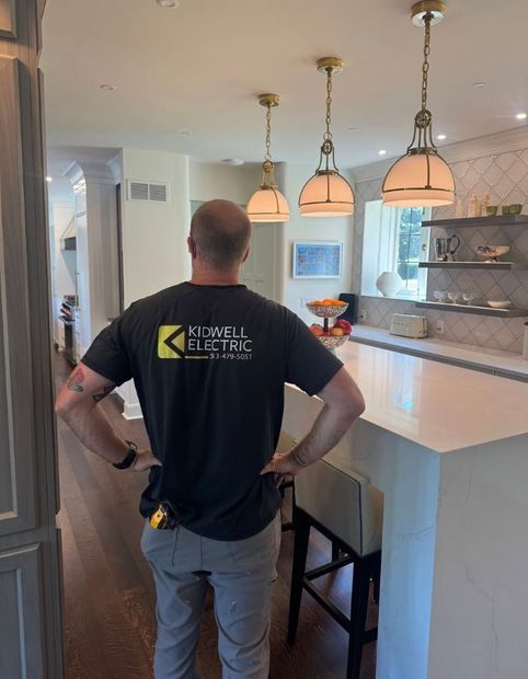 Electrician in black shirt standing in a bright kitchen with pendant lights and a marble island.