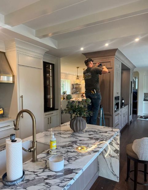 Person standing on a chair in a modern kitchen, installing cabinetry near a marble island.