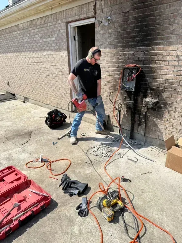 Worker near brick wall with tools and cables, inspecting an open utility panel outdoors.