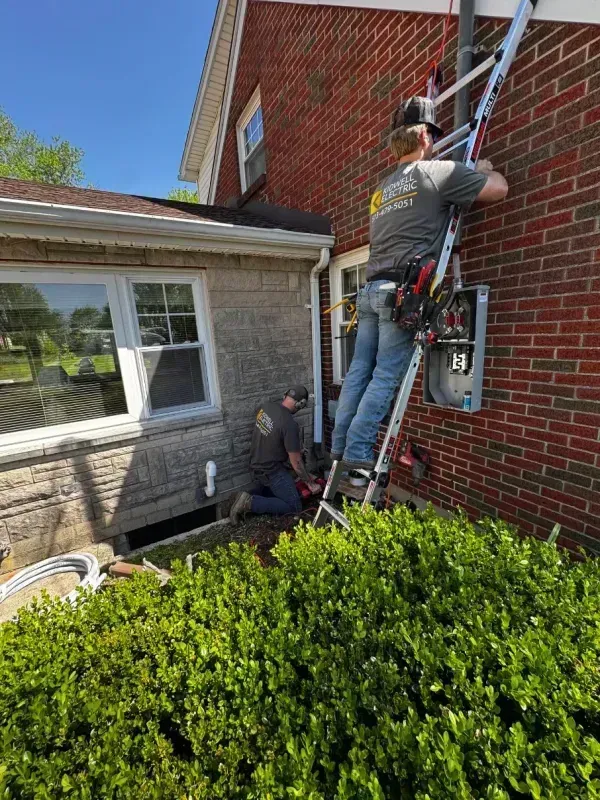 Worker on a ladder repairing a red brick wall beside a house with a hedge below