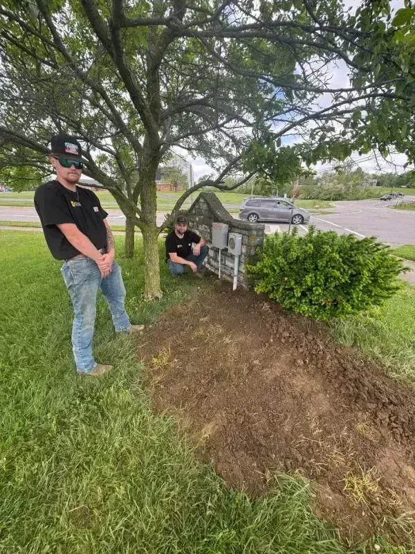 Two people stand by a tree and stone marker near a grassy roadside under cloudy skies.