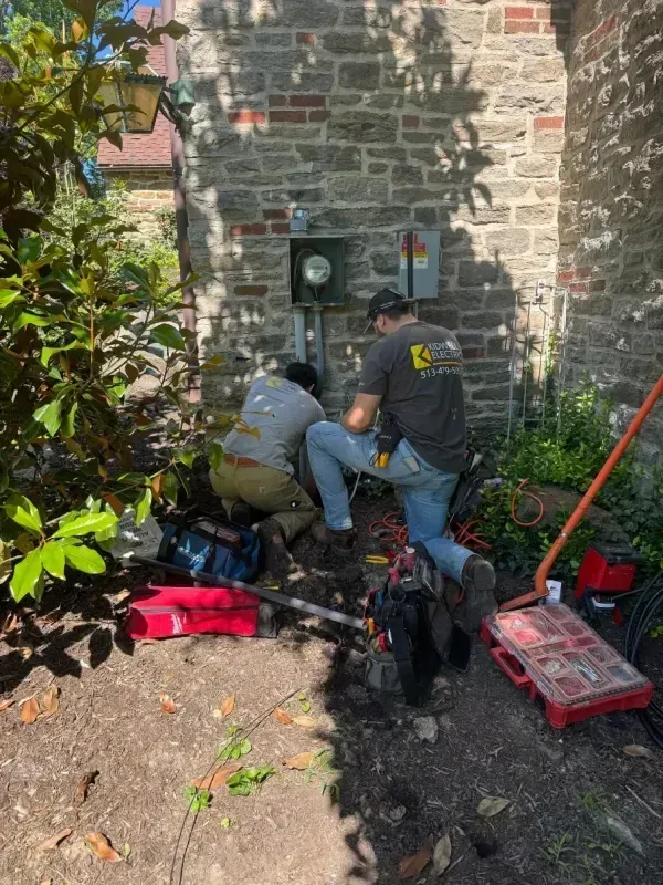 Two workers crouch by a stone wall, repairing an electrical box with tools and a red tool case nearby.