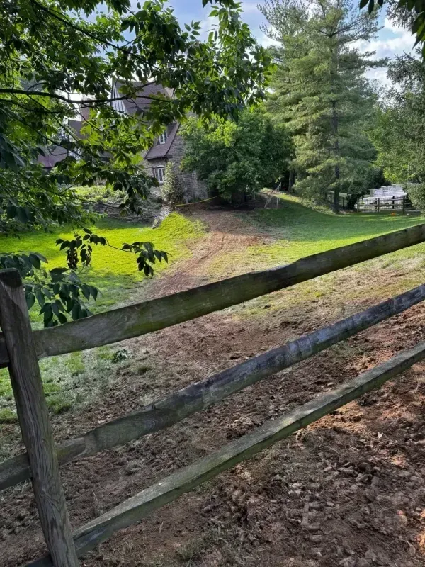 Wooden fence beside a grassy path leading to a house under leafy trees on a sunny day