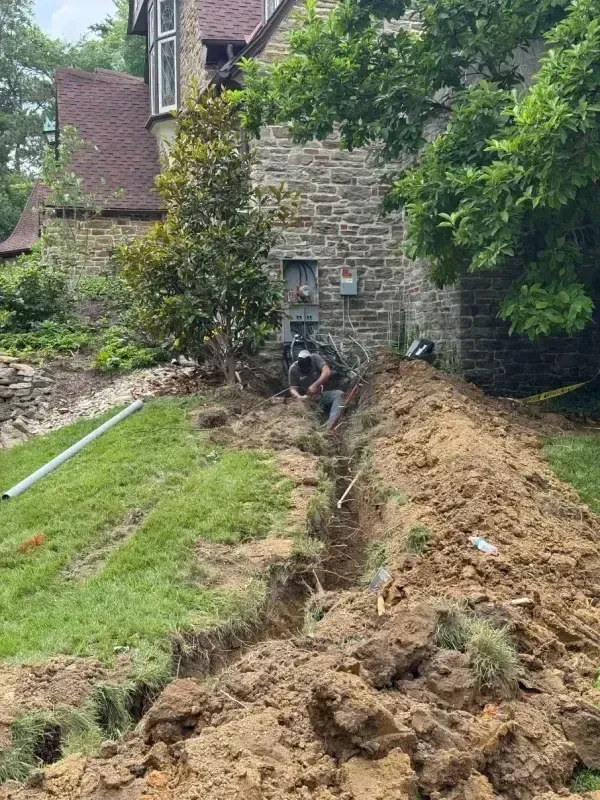 Backyard trench beside a stone house, with exposed soil and a worker digging near the wall