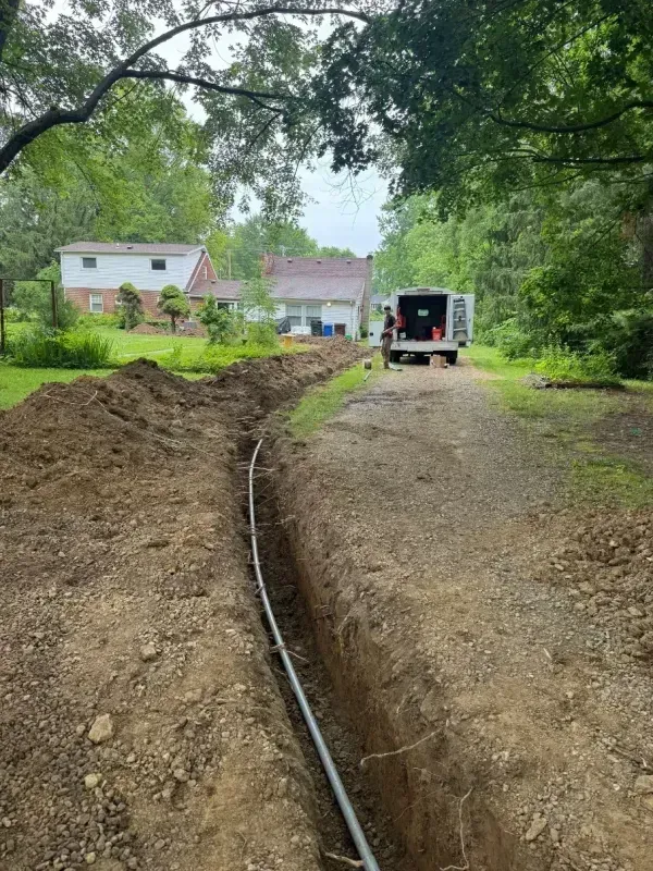 Trenched dirt driveway with a black pipe, leading to a work truck near houses and trees.