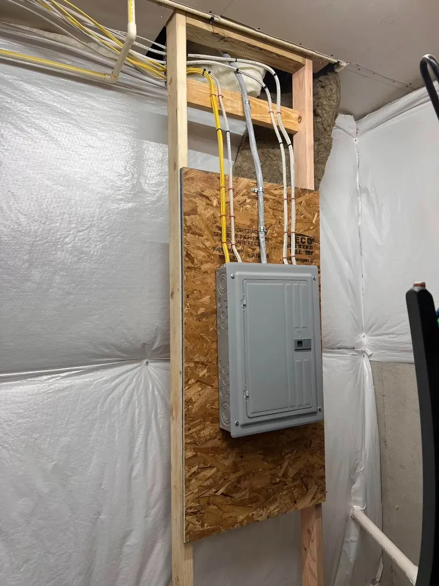 Electrical panel in an unfinished wall, with exposed insulation and conduit in a utility room.
