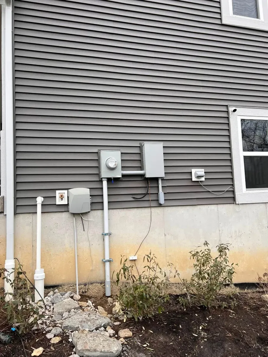 Gray house siding with utility boxes and pipes above a stained foundation wall with weeds and debris