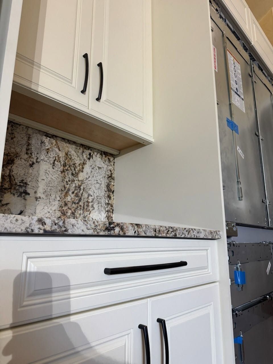 Kitchen cabinets with white drawers, speckled countertop, and exposed metal fridge area under construction