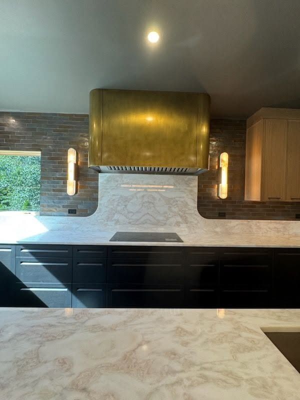 Modern kitchen with black cabinets, brass range hood, marble backsplash, and patterned countertop.