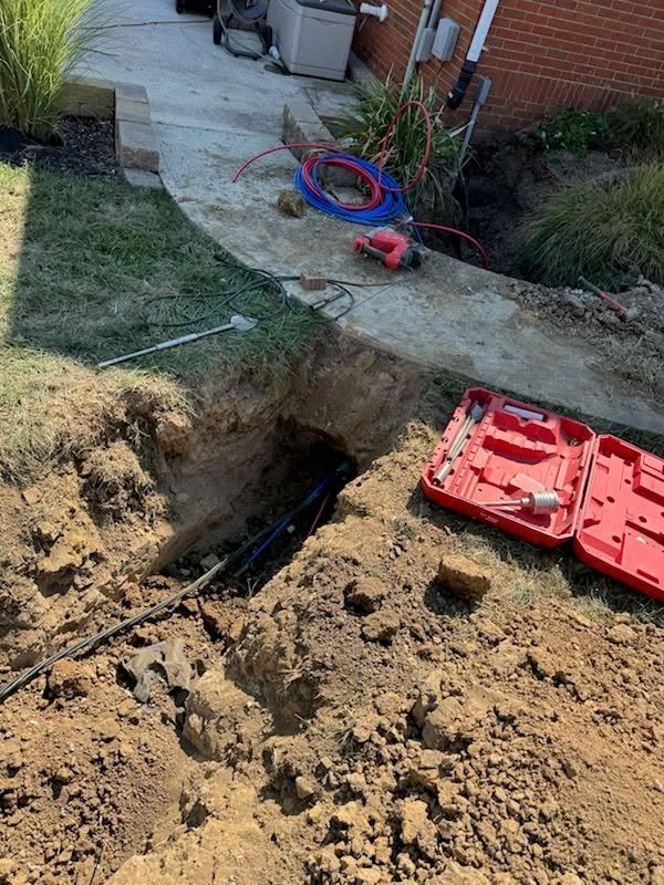 Backyard trench excavation with exposed pipes, dirt pile, and red tool case near a sidewalk