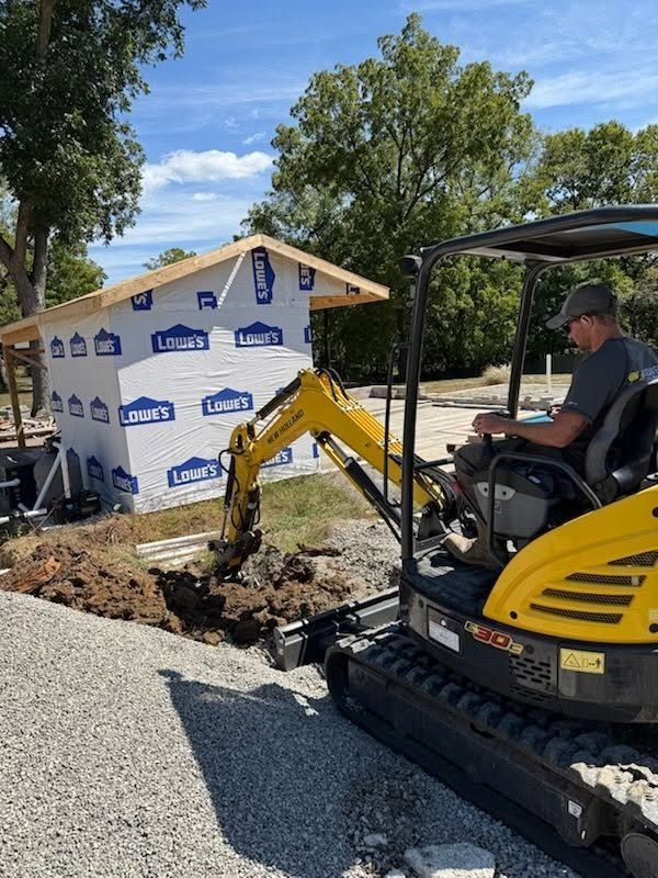 Mini excavator digging beside a house under construction, with gravel and a worker operating it.