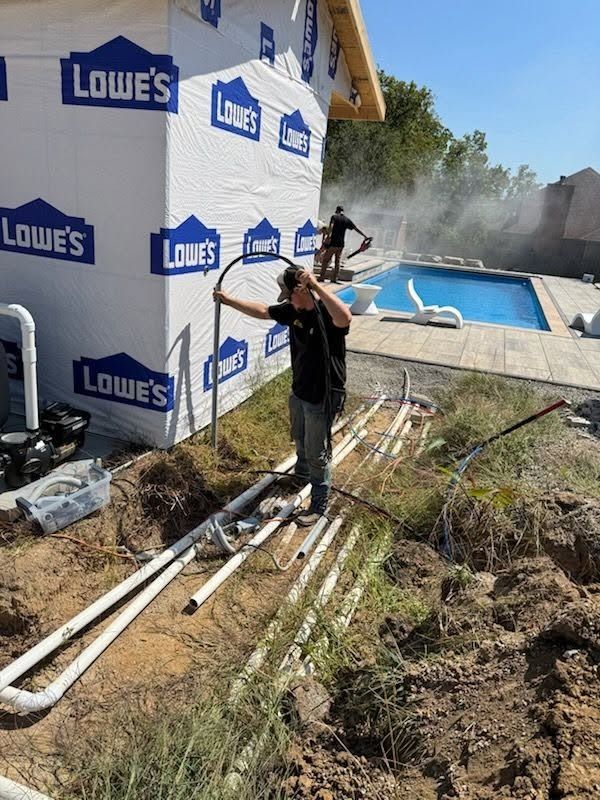 Worker on a construction site beside a Lowe's-wrapped house and a blue pool, moving debris with dust in the air
