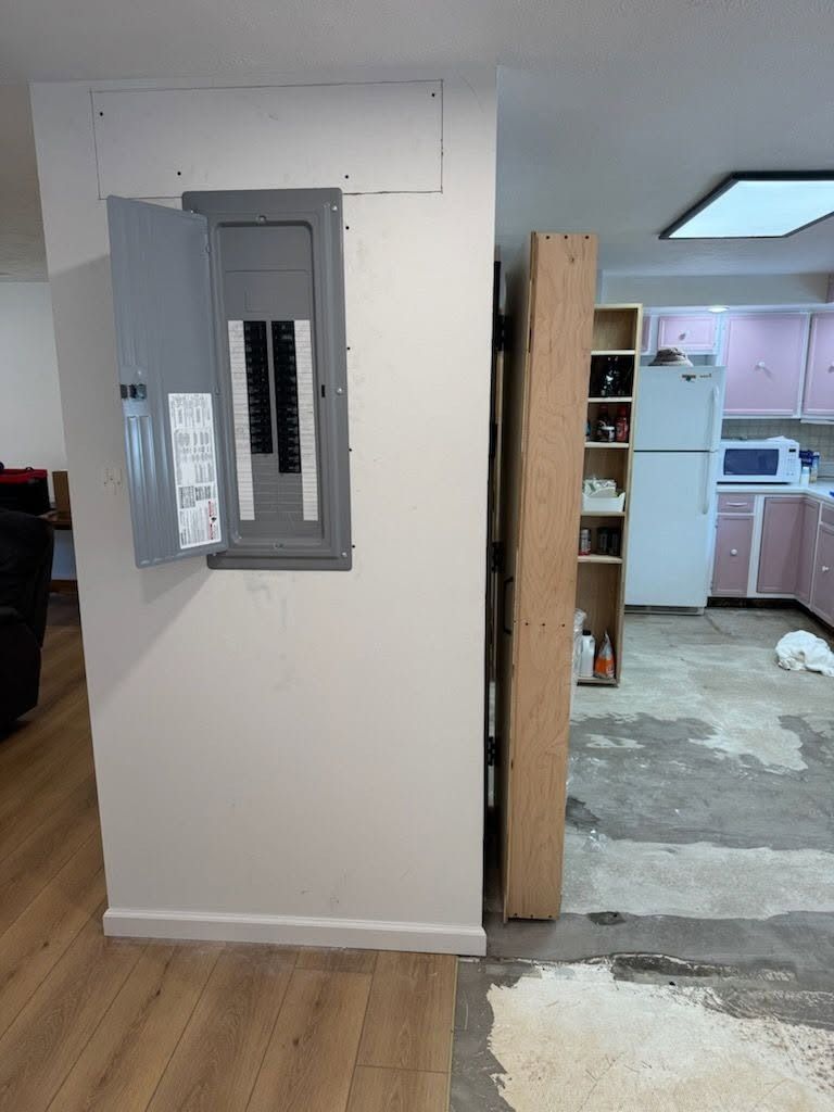 Interior hallway with open electrical panel on a white wall, leading into a damaged kitchen area with exposed floor.