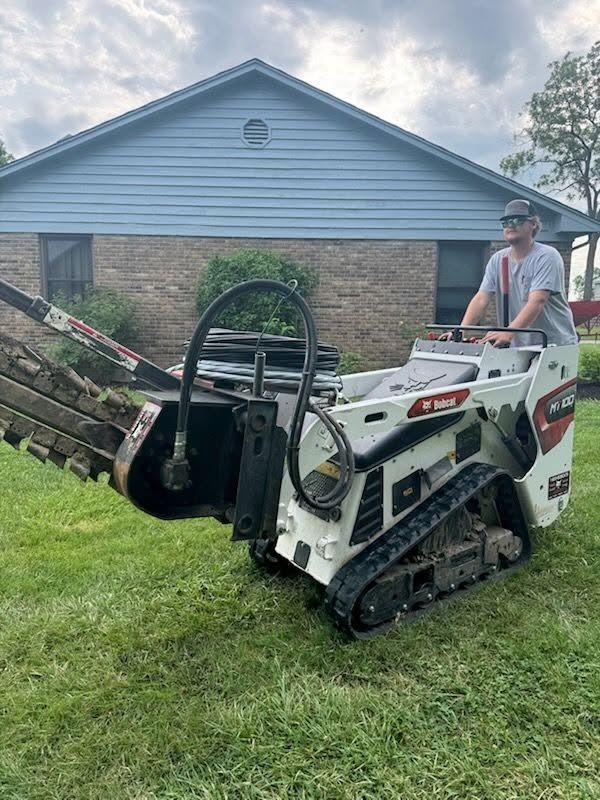 Man on a white skid steer lifting a metal trailer in a grassy yard beside a house