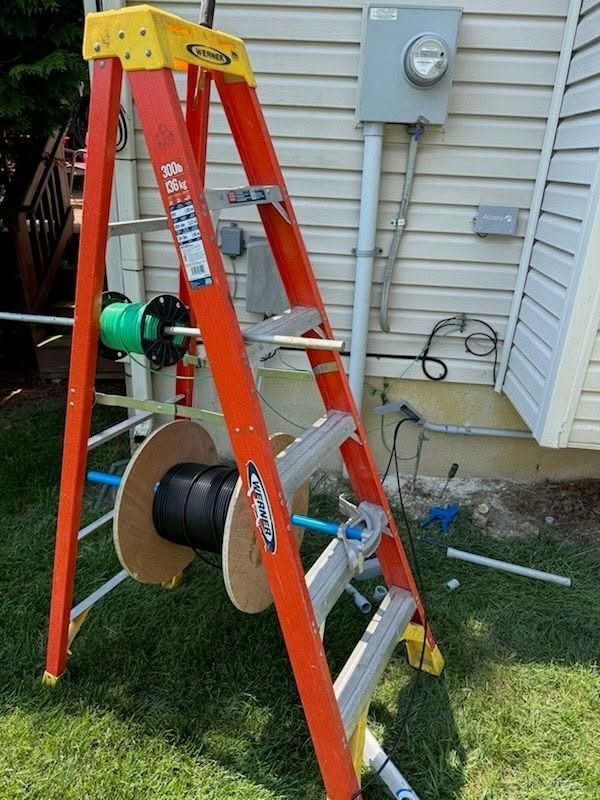 Orange ladder with cable spools in a yard beside a house and utility meter.