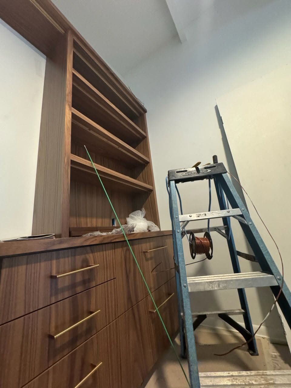 Wood cabinets under construction with a ladder, tools, and a green cable in a narrow room.