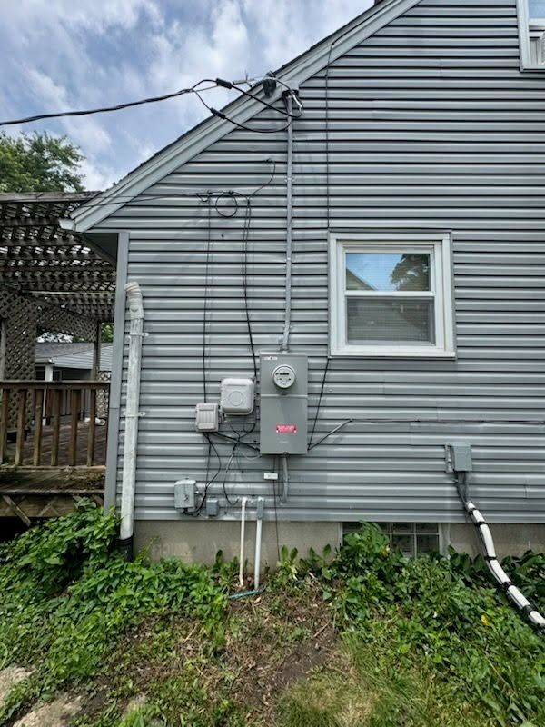 Gray house exterior with utility meters, cables, and a small window above a grassy slope.