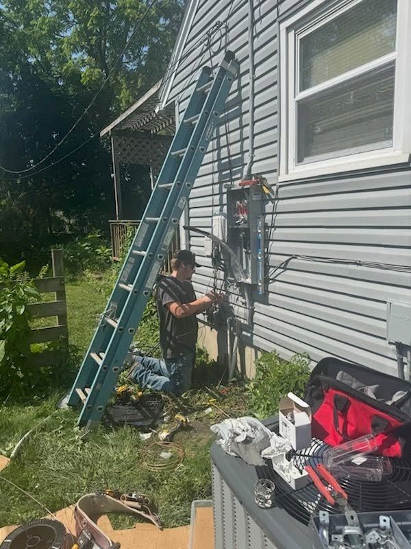Person on a ladder repairs house siding beside a window, with tools and supplies on the ground.
