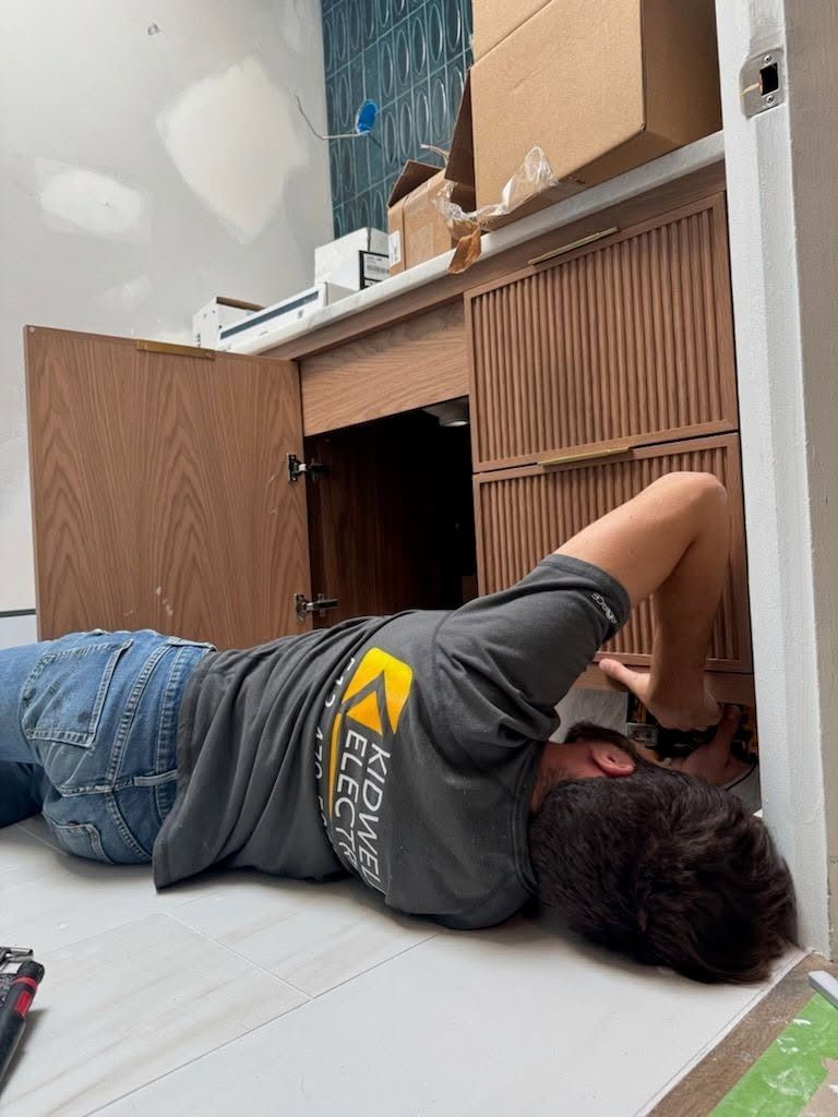 Person lying on the floor reaching into a wooden cabinet under a sink.