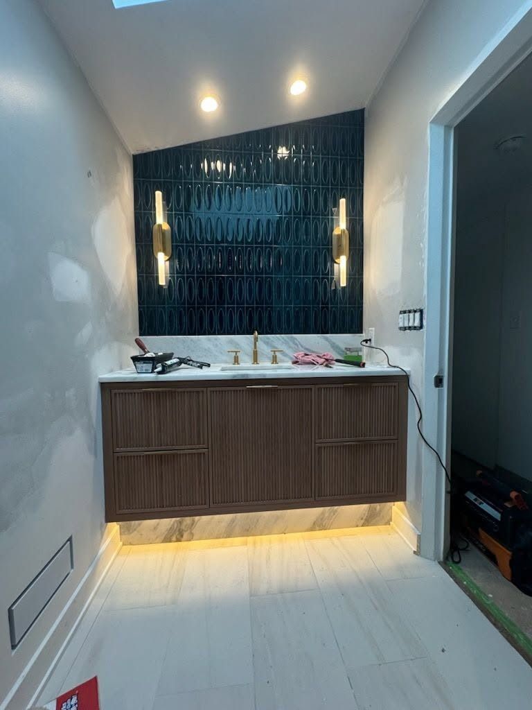 Modern bathroom vanity with wood cabinet, white countertop, dark textured backsplash, and warm under-cabinet lighting