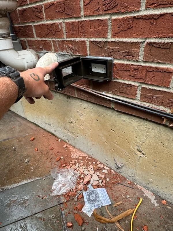 A hand points to an open wall vent in a brick exterior wall with concrete below.