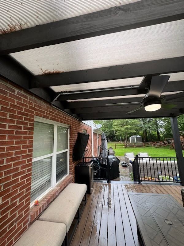 Covered brick patio with wet wooden deck, ceiling fan, bench, grill, and backyard gate.