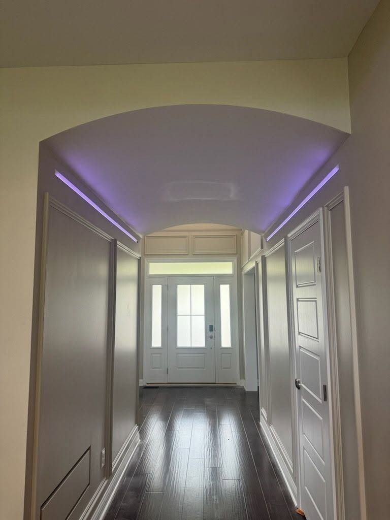 Narrow hallway with white doors, mirrored walls, and purple LED ceiling lights leading to a bright window.