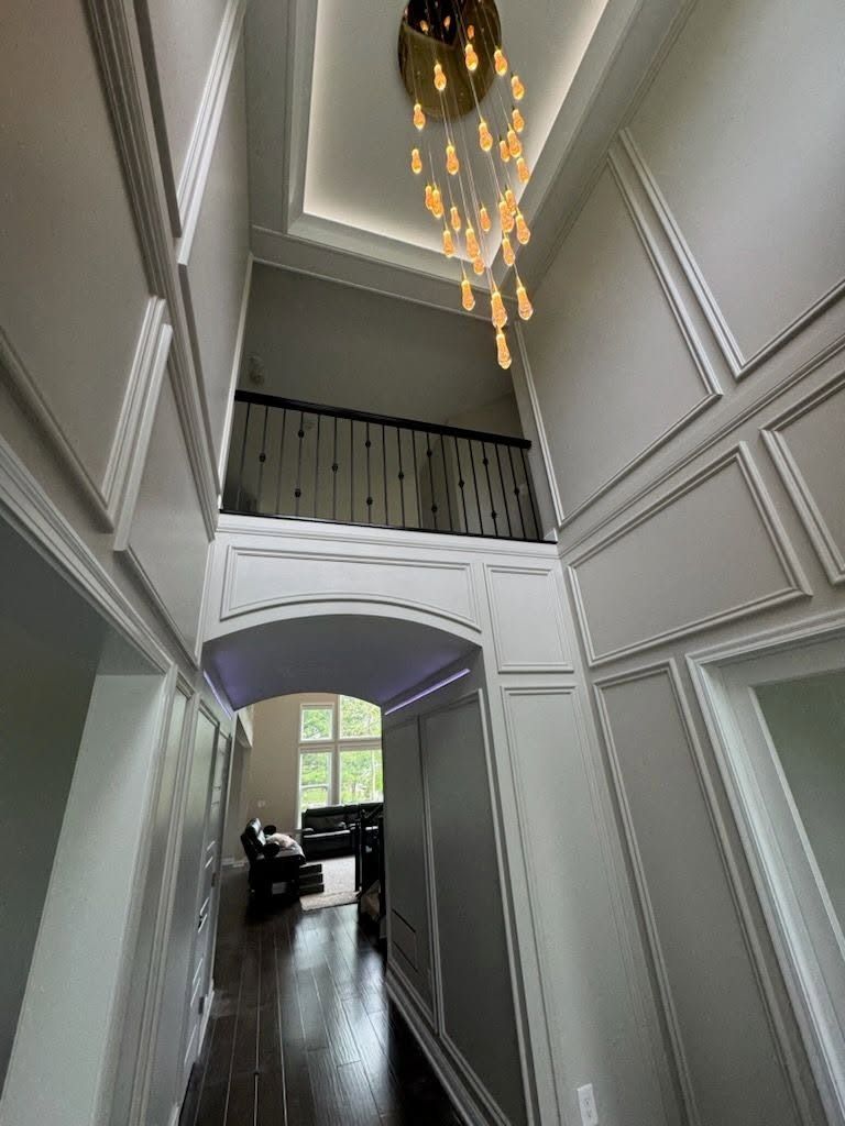 Tall white foyer with paneled walls, balcony railing, chandelier, and dark hardwood floor.
