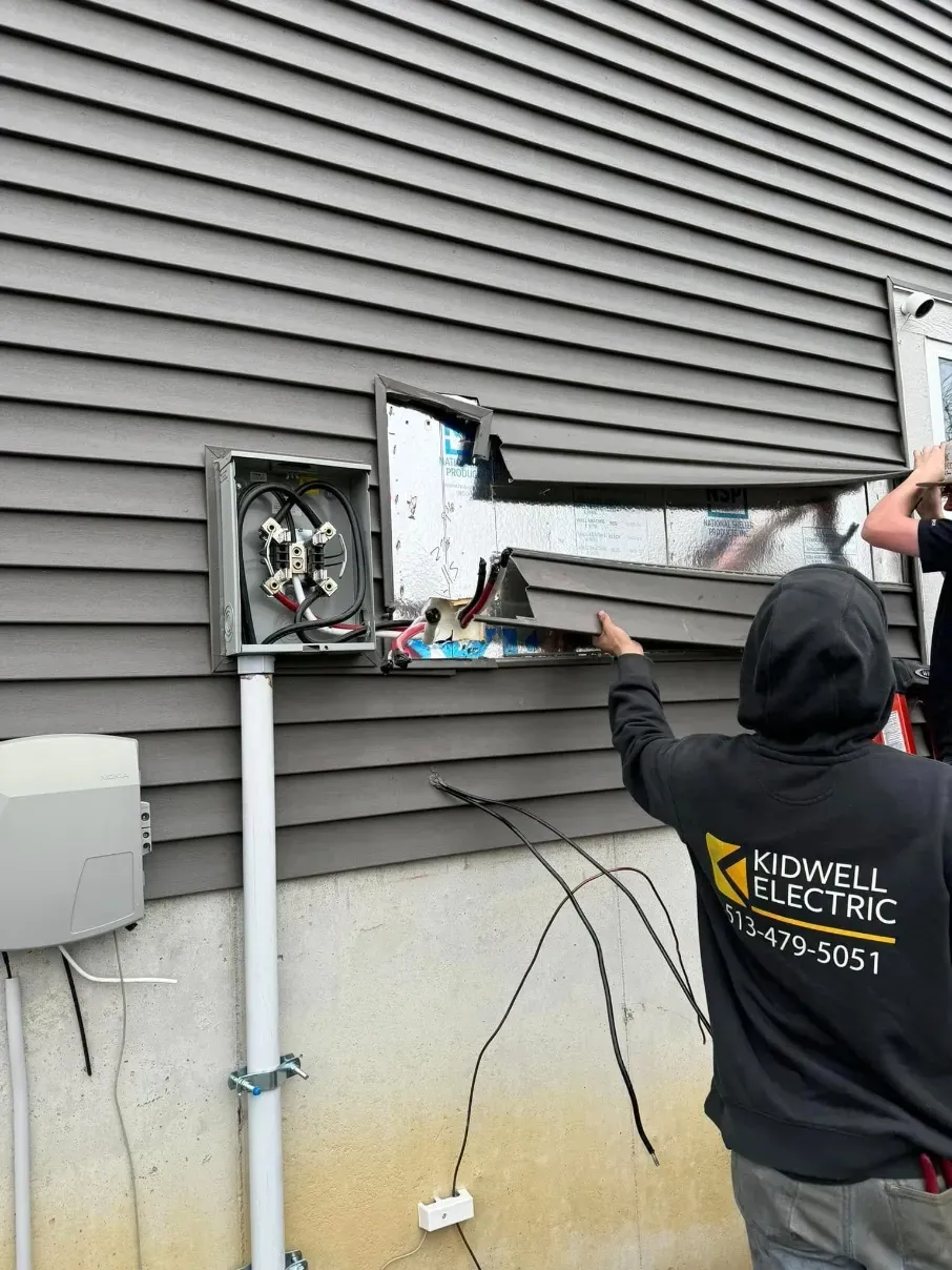 Person inspecting damaged siding on a house with exposed wiring and an open wall panel.