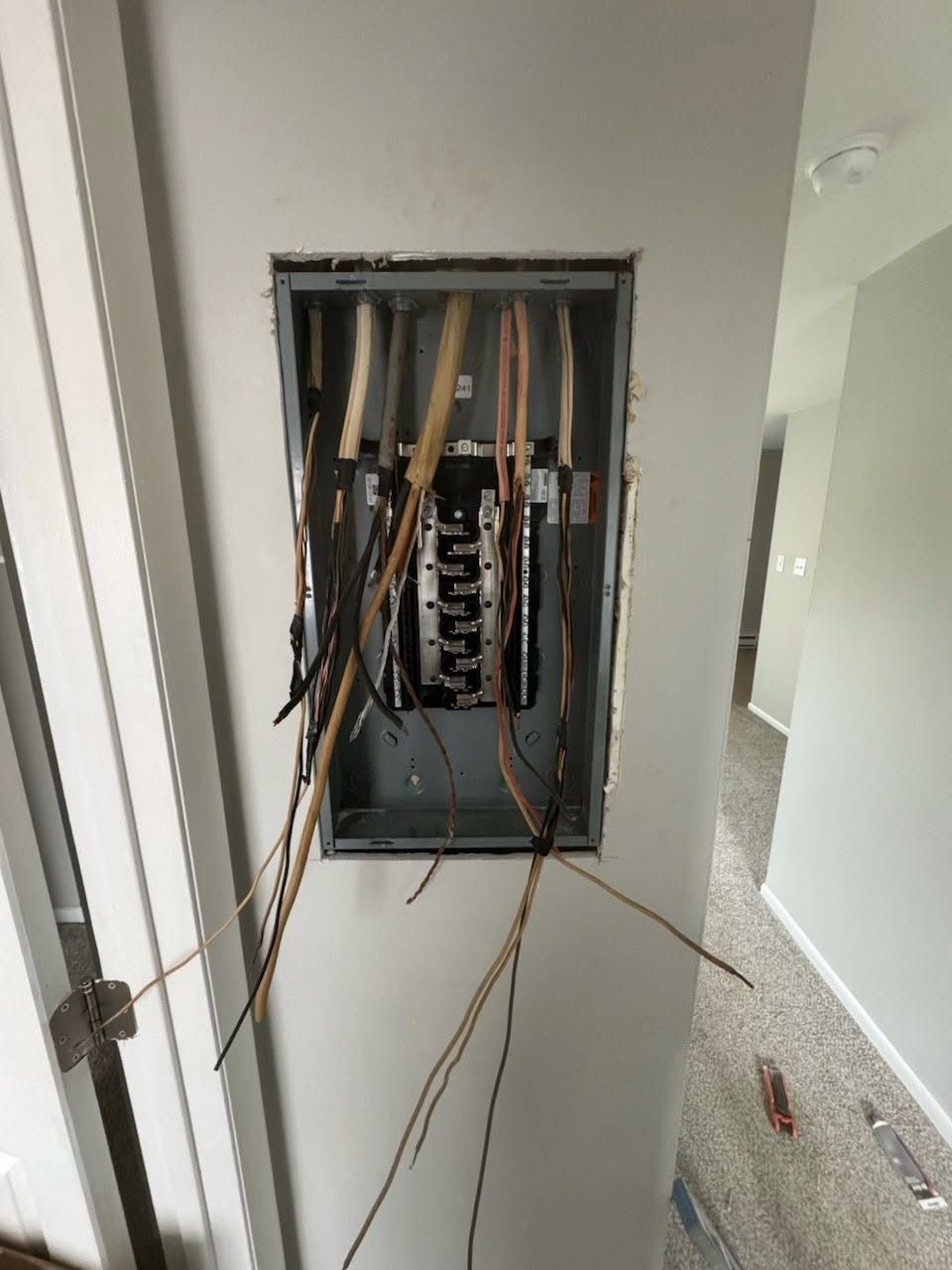 Open electrical panel with exposed wiring in a hallway wall opening.