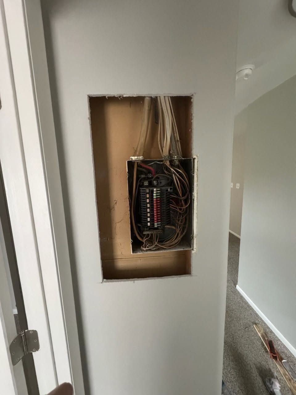 Open wall electrical panel with exposed wires and circuit breakers in a hallway