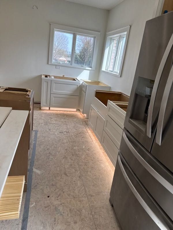 Narrow unfinished kitchen with white cabinets, stainless fridge, and windows at the far end.