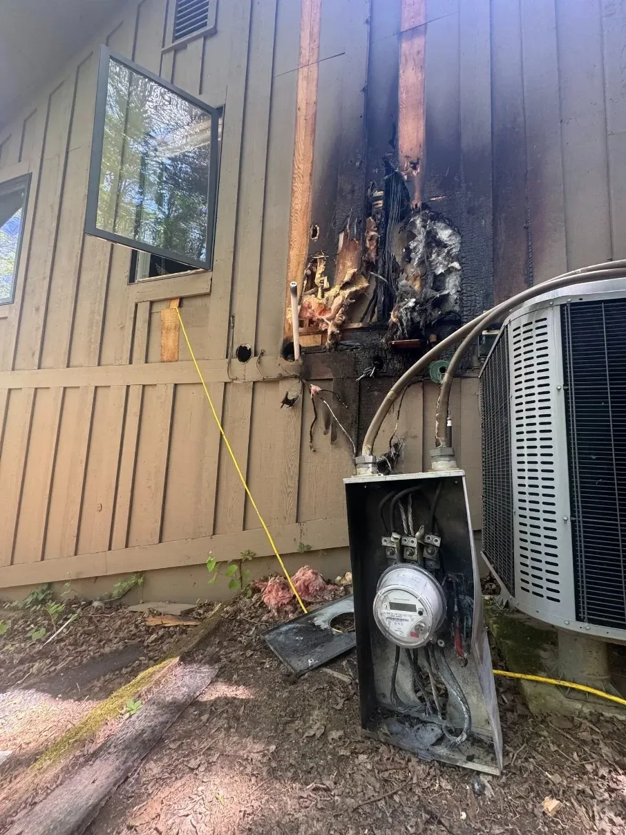 Charred exterior wall and utility meter after a fire, with melted siding and an air conditioner nearby