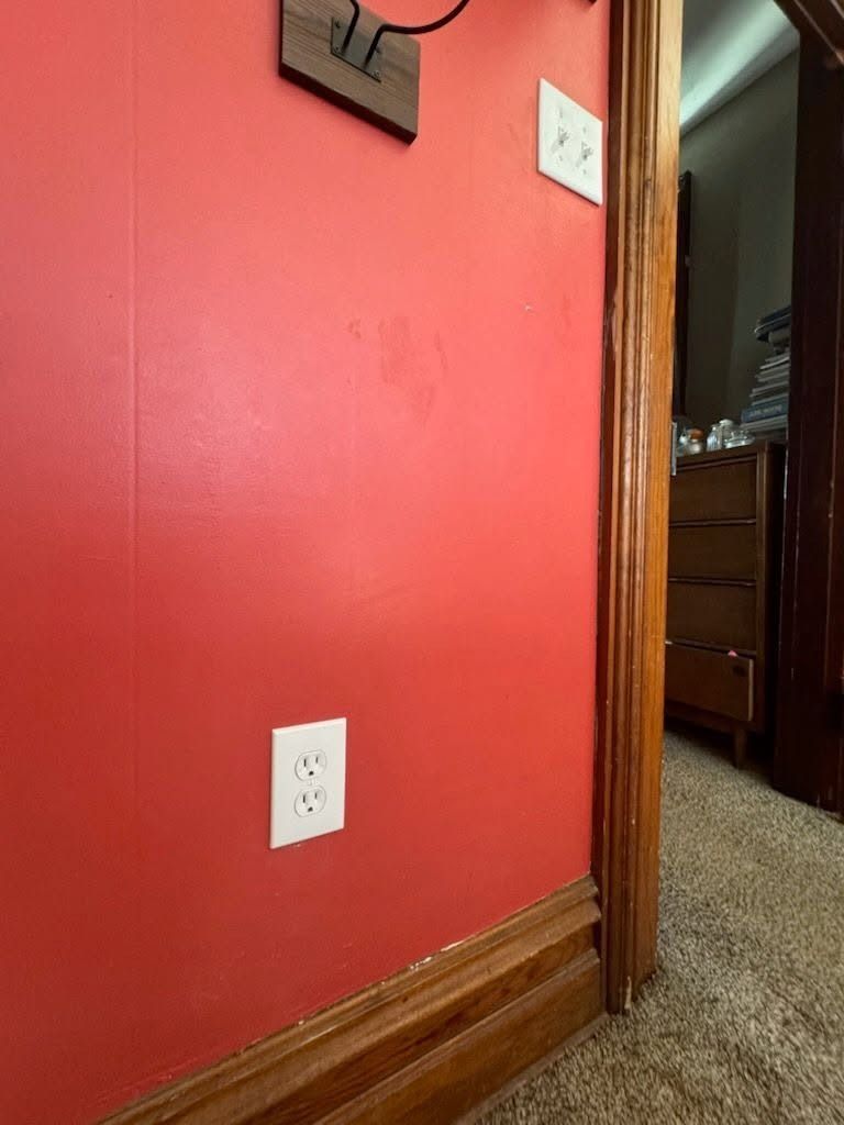 Red hallway wall with a light switch and wooden trim beside a doorway leading into a room.