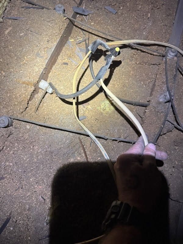 Hands holding electrical cables over a dusty floor with exposed wiring and a flashlight beam