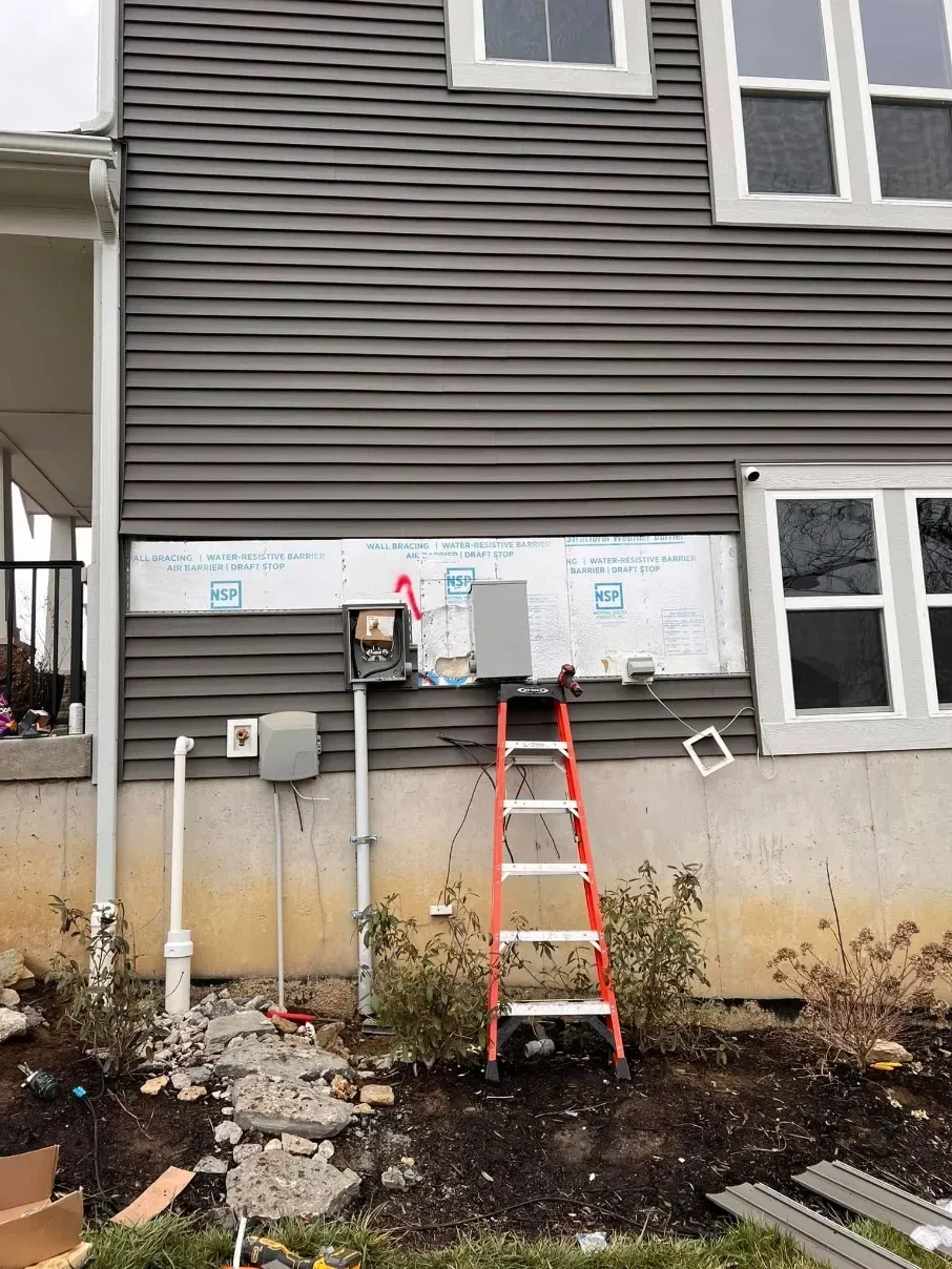 House exterior with utility meters and pipes, orange ladder against wall, and exposed foundation work below windows