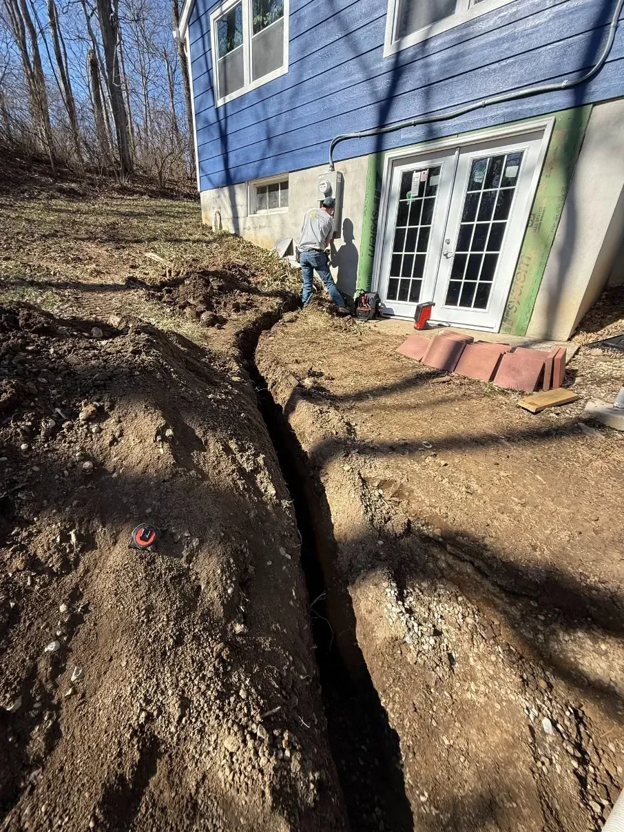 Excavated trench beside a blue house with French doors and a sloped dirt yard