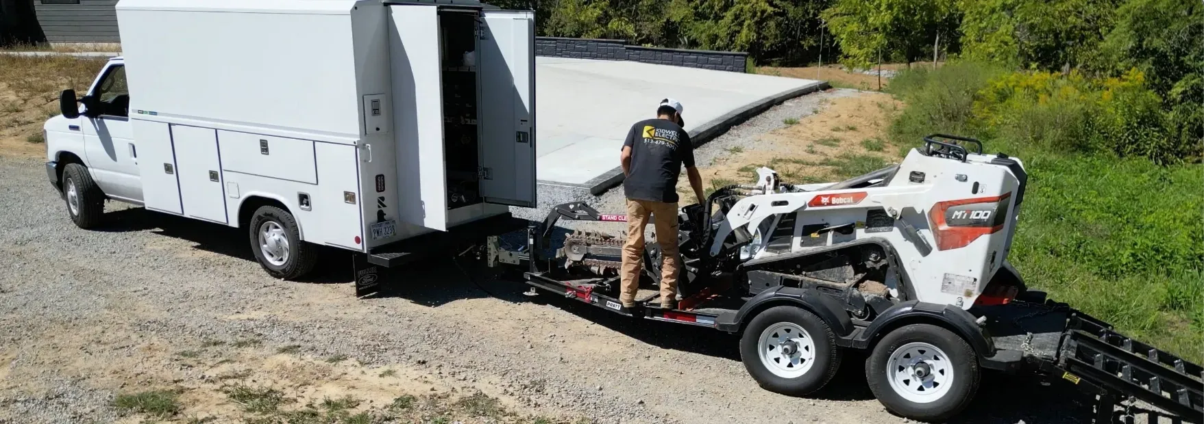 White utility truck with ramps and a man loading a white skid steer on a gravel roadside.