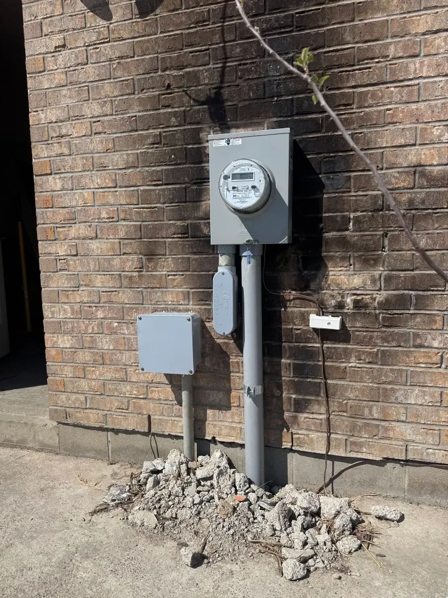 Electric meter and utility boxes on a brick wall with exposed conduit and broken concrete below.