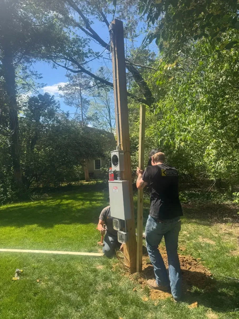 Two people work beside a tall wooden utility pole in a grassy yard with trees and a white electrical box.