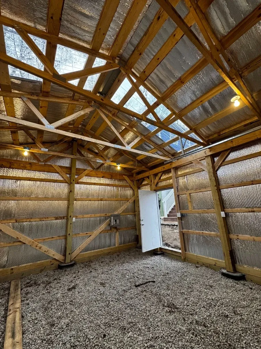 Interior of a small unfinished shed with wooden trusses, insulation, gravel floor, and an open white door.