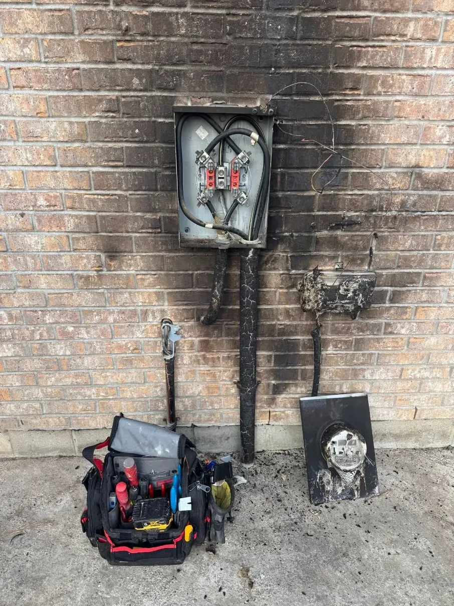 Exposed electrical panel on brick wall with tools and equipment on the ground below.