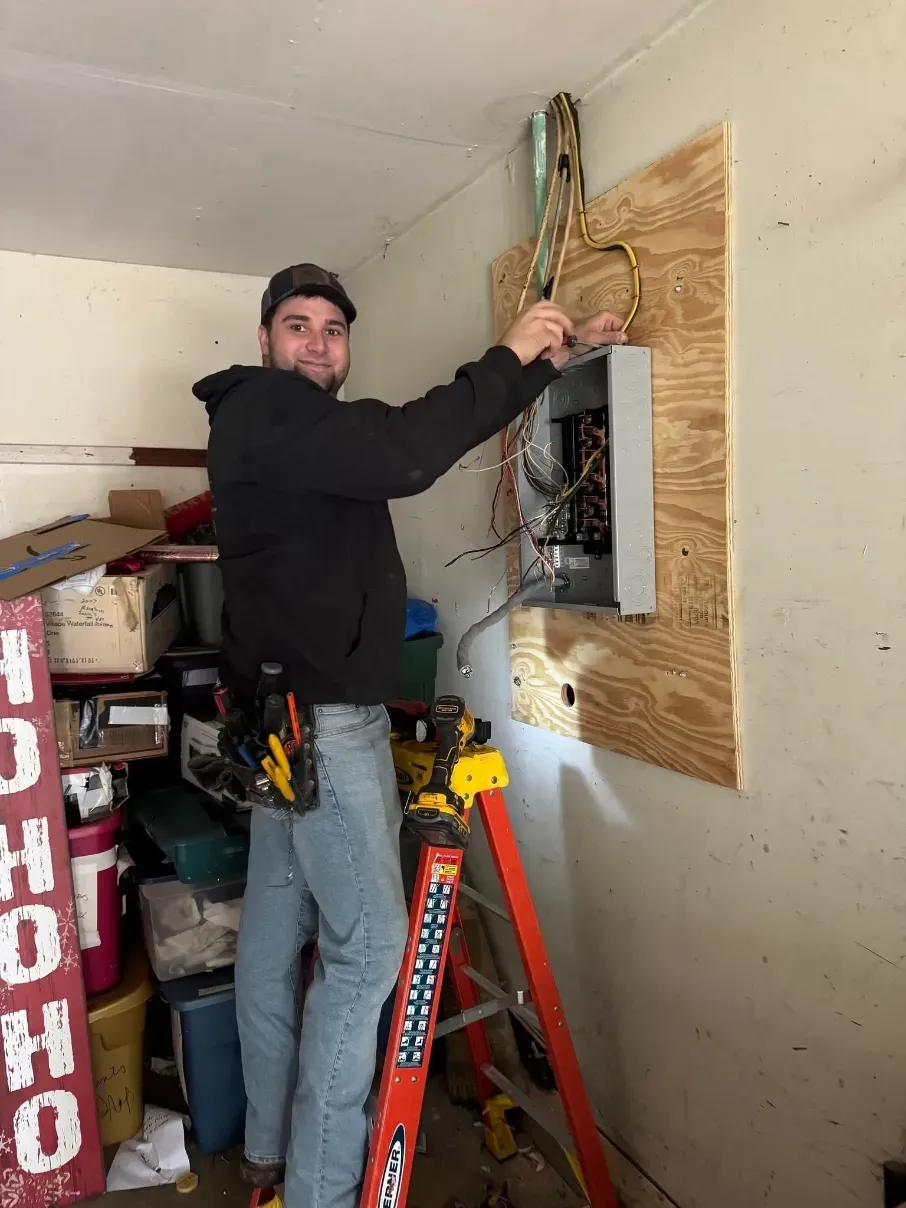 Technician in black jacket wiring an electrical panel in a small unfinished room.