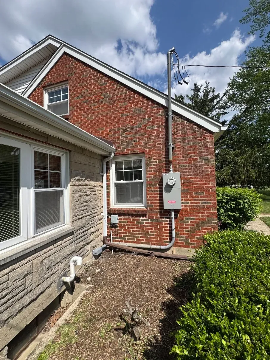 Brick house exterior with utility meter and white window beside a trimmed hedge under a blue sky
