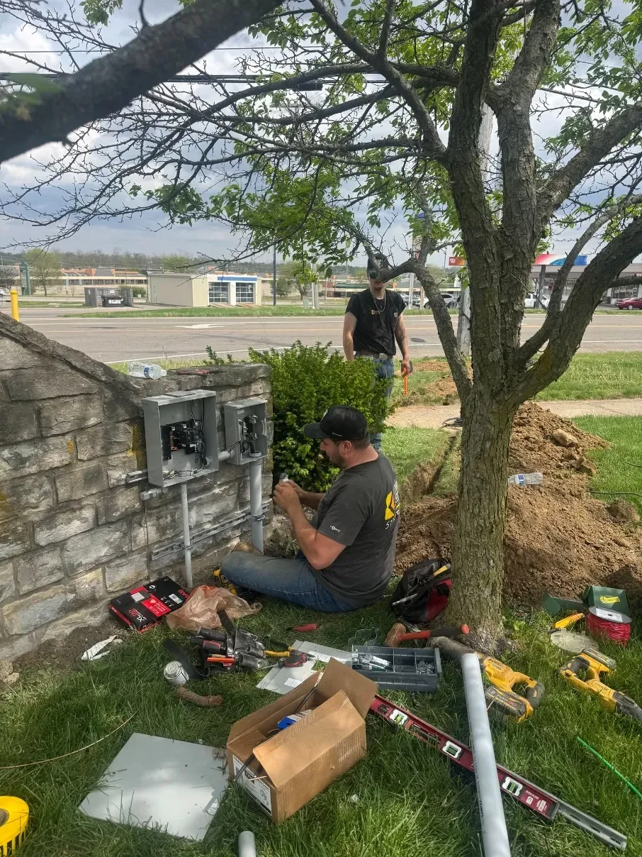 Technician kneels by an outdoor electrical box under a tree, with tools and cables spread on the grass.