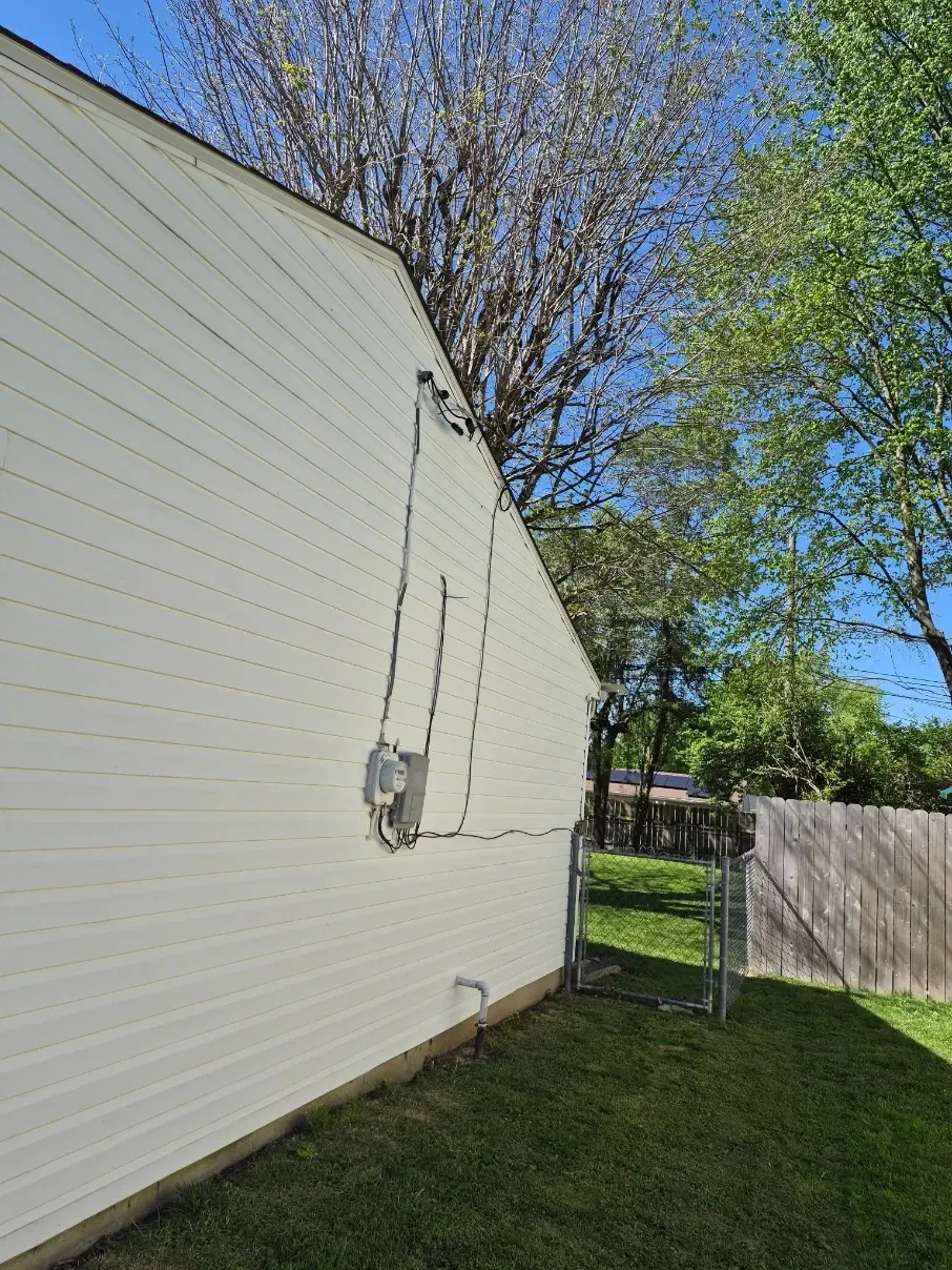 Side of a beige house with electrical wiring, grassy yard, wooden fence, and trees under a blue sky.